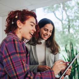 A young trendy businesswoman is holding a tablet and pointing at it while giving a positive feedback to her colleague for a report. Associate evaluating work while pointing at the tablet at office.